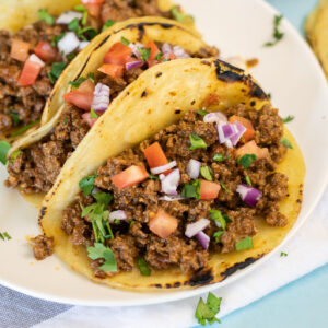 Close up of a ground beef taco in a corn tortilla filled with crockpot taco meat topped with pico de gallo on a plate with two other tacos.