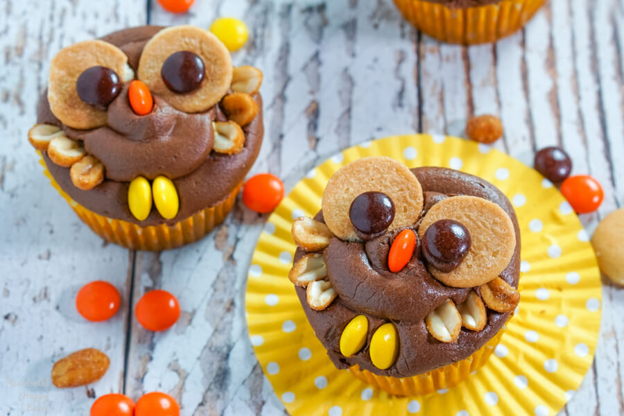 Two peanut butter chocolate owl cupcakes sit on a yellow cupcake wrappers surrounded by Reese's Pieces candy.