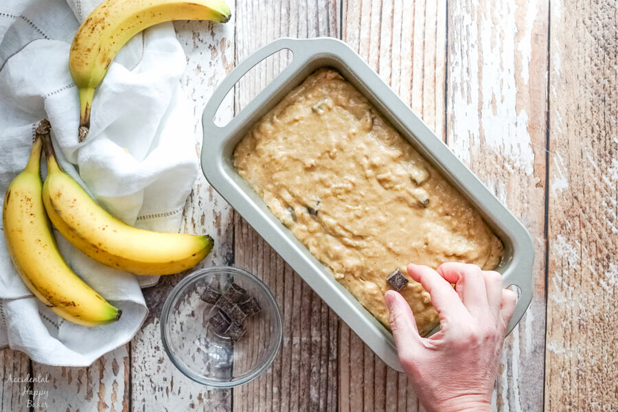 A loaf pan full of banana bread batter as more chocolate chunks are added to the top. 