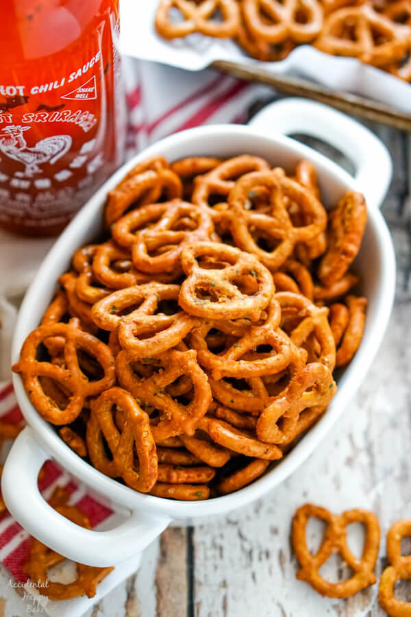 A while bowl full of spicy ranch pretzels next to a bottle of sriracha sauce.