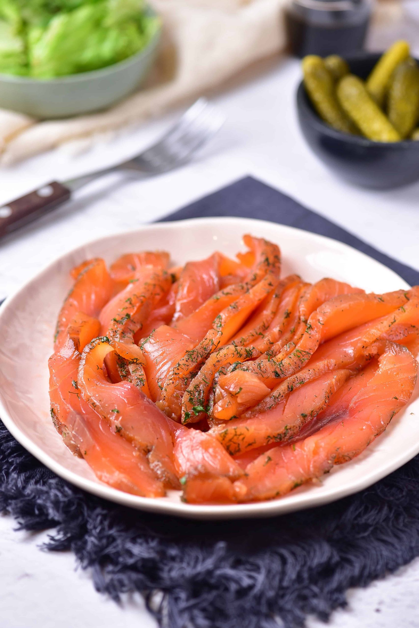 A plate of sliced gravlax salmon garnished with dill is placed on a dark textured cloth. In the background, a bowl of pickles, a bowl of lettuce, a fork, and a knife are visible, all set on a light-colored tabletop—an easy recipe for an elegant meal.