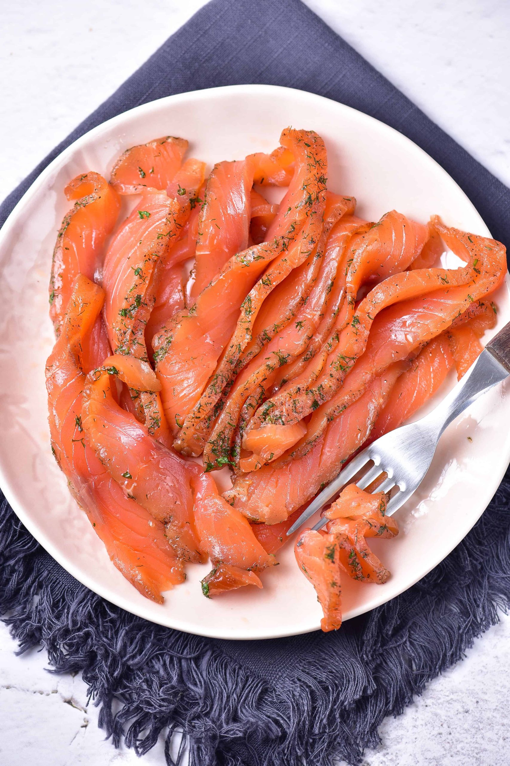 A white plate of delicately sliced gravlax salmon, garnished with fresh dill, sits on a dark napkin. A fork cradles a small portion of the easy gravlax, ready to enjoy. The backdrop is a textured white surface.