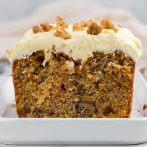 A sliced loaf of carrot cake with cream cheese frosting and chopped walnuts on top, displayed on a white dish.