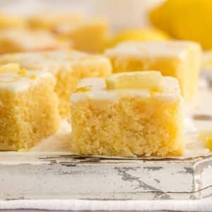 Close-up of lemon brownies topped with glaze and small lemon pieces, arranged on a rustic white surface.
