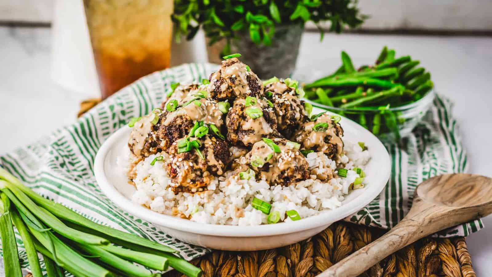 A bowl of rice topped with meatballs in a creamy sauce, garnished with chopped green onions. It is placed on a striped cloth, accompanied by a wooden spoon and a bowl of green beans. A beverage is visible in the background.