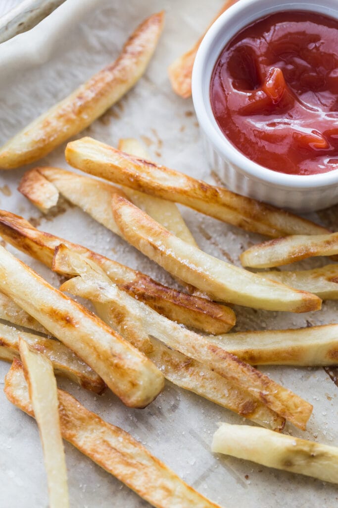 Overhead photo of golden brown homemade baked French Fries on a parchment lined sheet pan. There is a white ramekin of ketchup on the corner of the pan. 