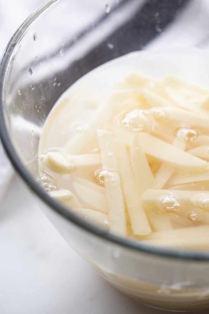 Raw thin cut potatoes soaking in water in a glass bowl. 