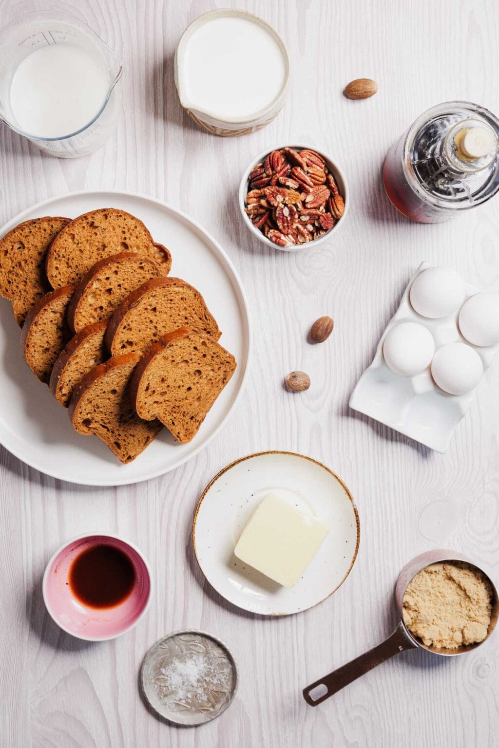 Slices of bread on a plate, accompanied by ingredients like milk, nuts, syrup, eggs, butter, vanilla extract, brown sugar, and a bowl of spices. Perfect for crafting a gluten-free French toast casserole on a light wooden surface.
