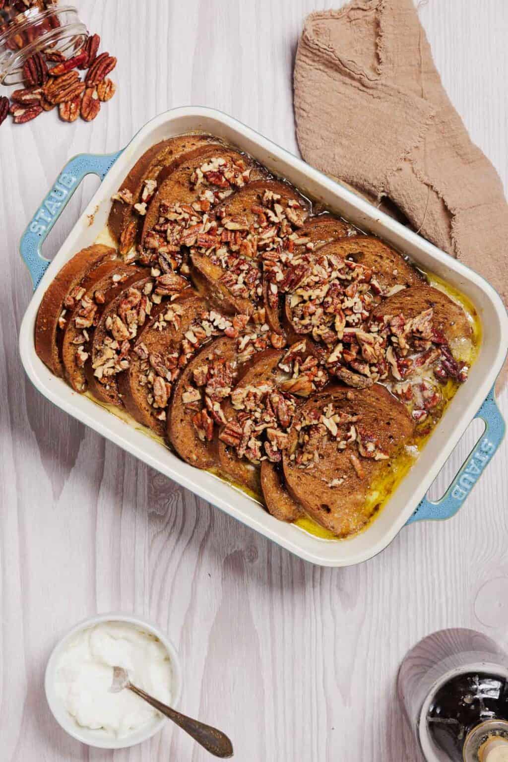 A baking dish contains slices of gluten-free French toast casserole topped with chopped pecans, nestled in a custard mixture. Nearby, a beige cloth, a glass of water, and a bowl with a spoon accompany the scene. A jar tipped over spills pecans onto the white wooden surface.