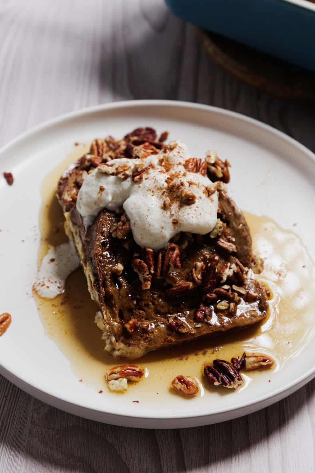 A plate of gluten-free French toast casserole topped with pecans, whipped cream, and a dusting of cinnamon. Syrup is elegantly drizzled around. The pristine white plate sits invitingly on a rustic wooden surface.