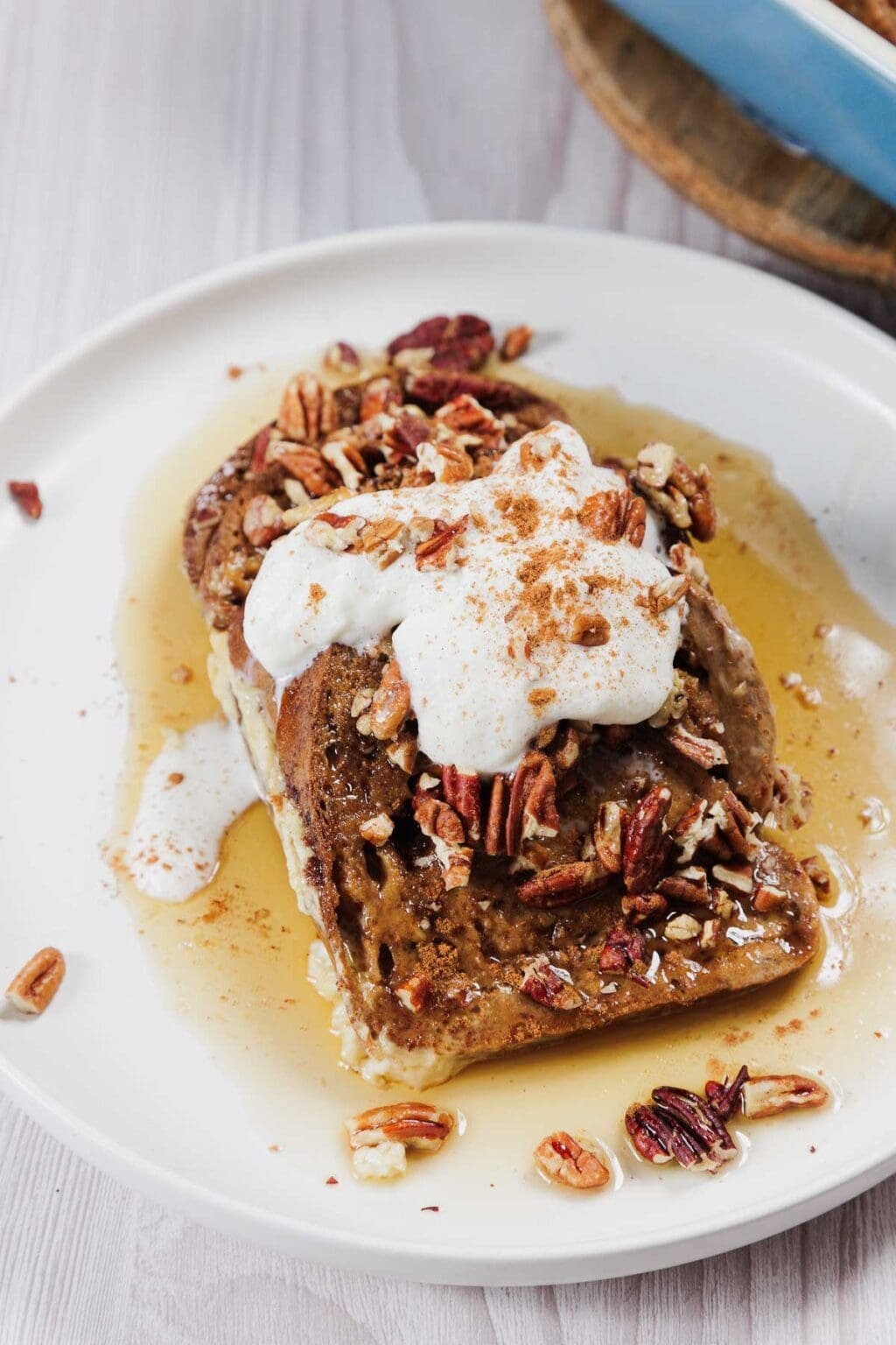 A plate with slices of gluten-free French toast casserole topped with whipped cream, chopped pecans, and a sprinkle of cinnamon. Syrup is drizzled over the toast, creating a small pool on the light-colored wooden surface beneath.