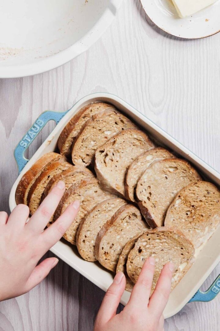 Two hands arranging slices of whole-grain bread in a blue baking dish, preparing the perfect base for a gluten-free French toast casserole on a light wood surface. A partial view of a white plate with butter is visible in the background, hinting at delicious flavors to come.