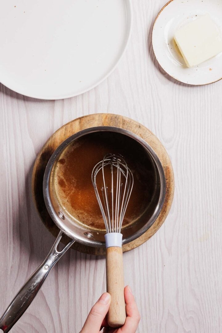 A hand skillfully wields a whisk over a saucepan filled with rich, brown sauce on a wooden board. Nearby, on a light wooden surface, rests the makings for gluten-free French toast casserole: a round plate and two squares of butter waiting to join the culinary creation.