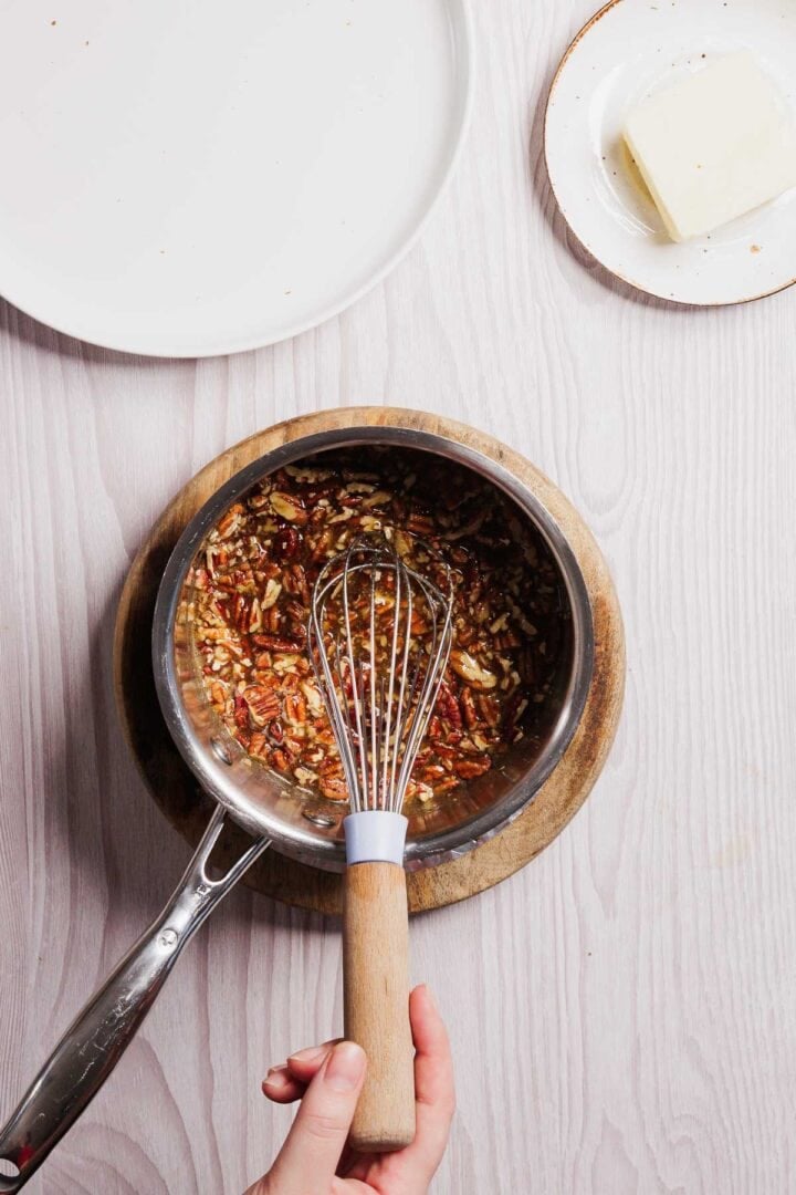 Top view of a saucepan on a wooden board containing mixed ingredients with pecans and a whisk, perfect for a gluten-free French toast casserole. A hand holds the whisk, while nearby, a small plate holds a block of butter. An empty white plate is partially visible above on the white wooden surface.