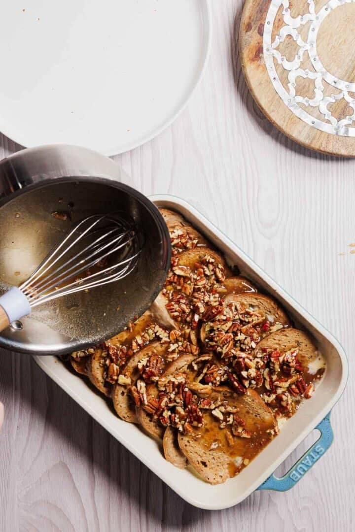 A baking dish filled with sliced bread, pecans, and syrup is being prepared for a gluten-free French toast casserole. A hand skillfully pours a liquid mixture over the contents from a pot with a whisk. Nearby, on the light wooden surface, rests a wooden board with an elegant decorative plate.