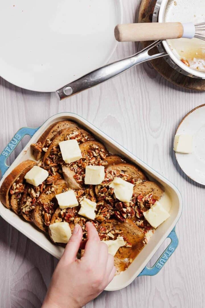 A hand places pats of butter on slices of bread topped with chopped pecans in a blue baking dish, assembling a delightful gluten-free French toast casserole. Nearby, a saucepan with a wooden handle and an empty white plate rest on the light wooden surface.