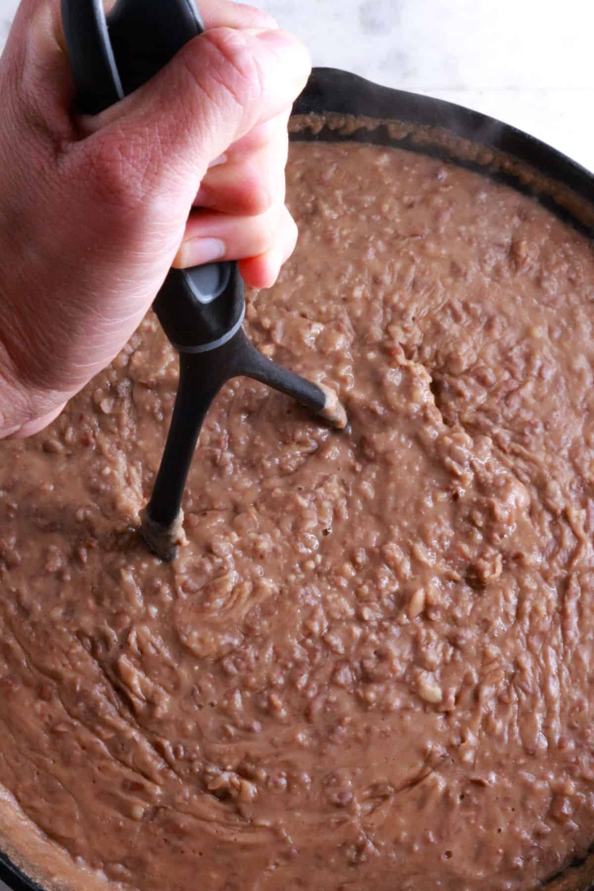 A hand on a potato masher mashing beans in a cast-iron skillet