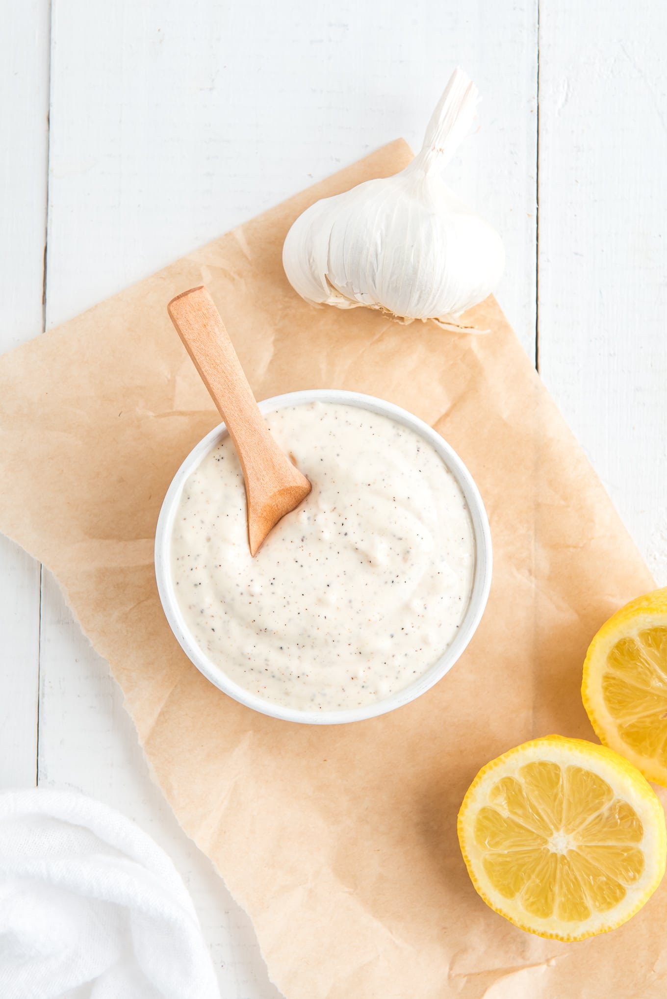 A bowl of aioli on the table on top of a piece of parchment with head of garlic and lemon slices to the side.