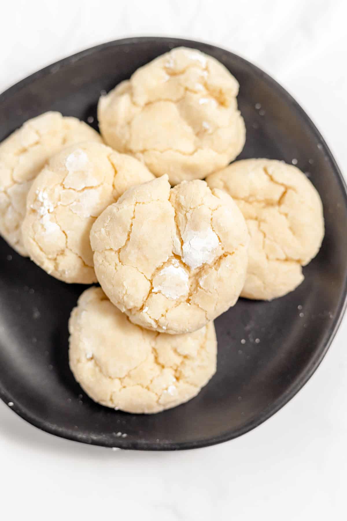 A black plate holds a stack of round, gooey cookies, their cracked surfaces dusted with powdered sugar on a pristine white surface.