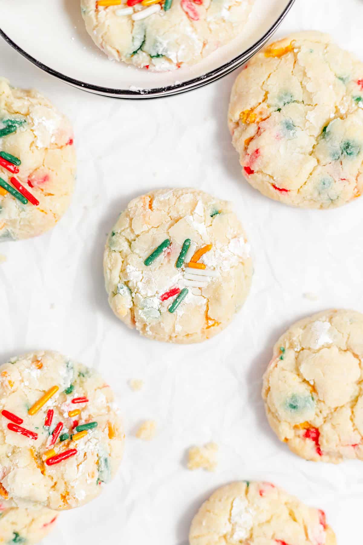 Gooey butter cookies adorned with vibrant sprinkles rest on crinkled parchment paper, with one temptingly perched on a plate.