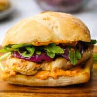 Closeup side view of a grilled chicken sandwich topped with grilled red onions and arugula on a wooden board.