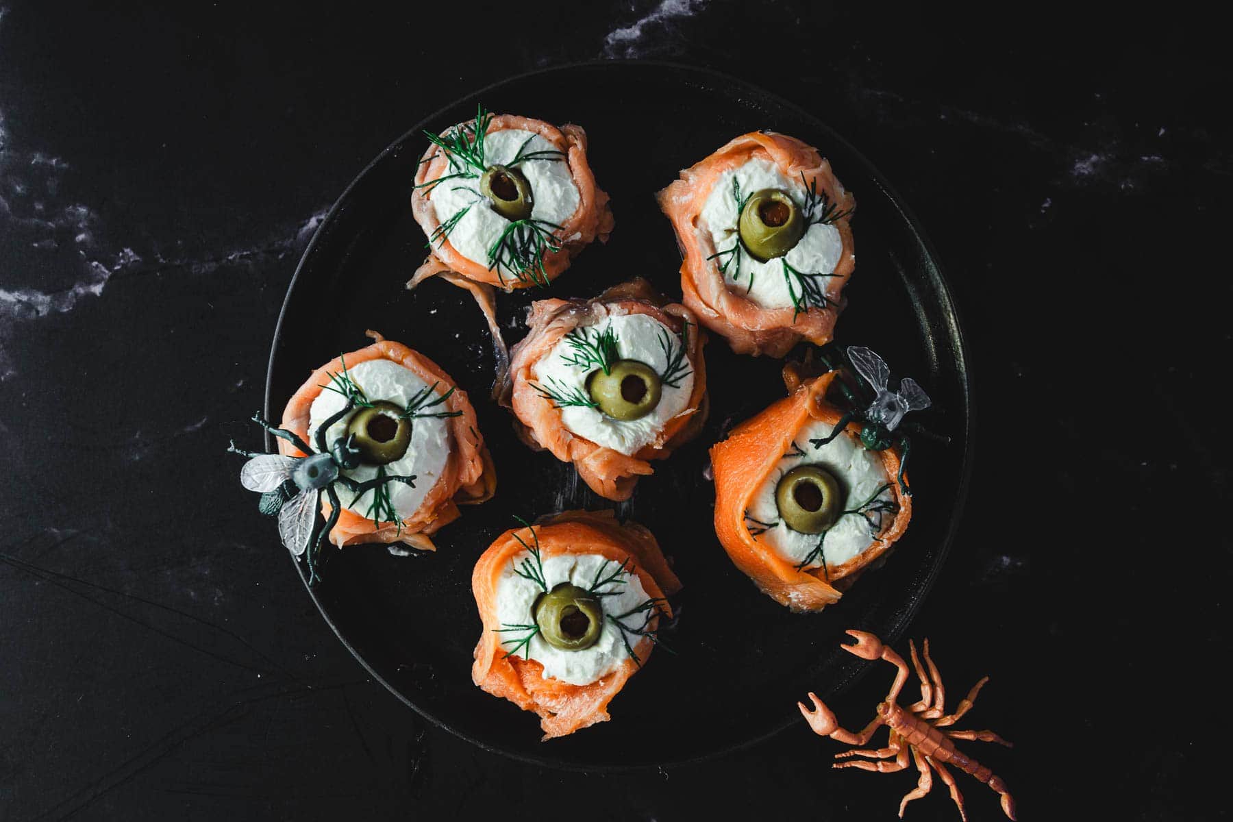 A black plate holds six creatively arranged appetizers resembling eyes, made from circular slices of salmon topped with cream cheese, a green olive slice, and a sprig of dill.