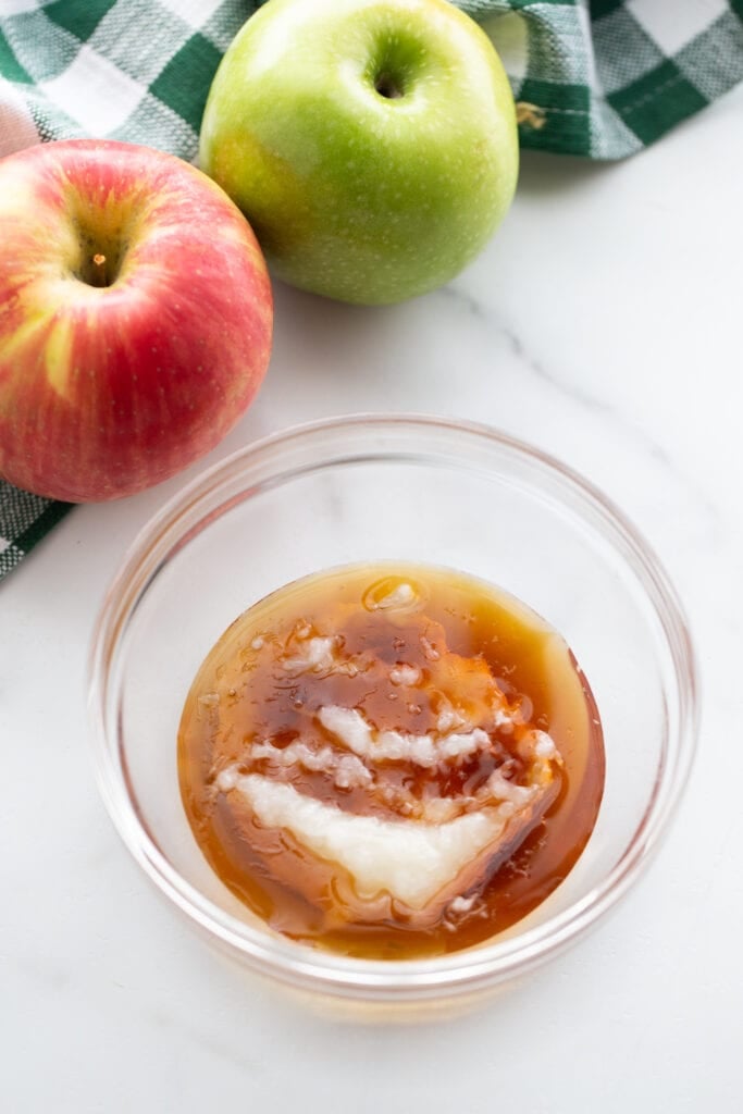 Small glass bowl of maple syrup and coconut oil, ready for the microwave to make caramel dip. On the side of the bowl are two whole apples.