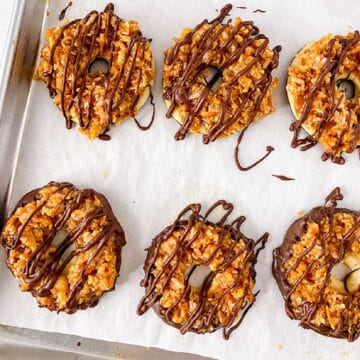 Six homemade Samoa cookies topped with caramel, coconut, and a chocolate drizzle sit on a parchment-lined baking sheet.