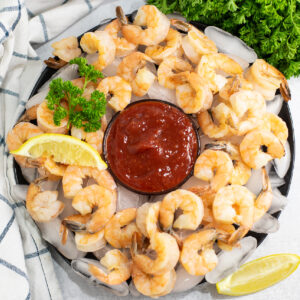 Overhead view of homemade shrimp cocktail on a plate of ice with a small black bowl of cocktail sauce in the center topped with a lemon wedge and a sprig of parsley.
