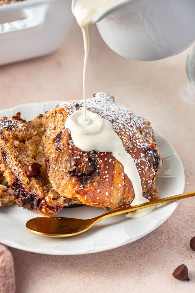 A small jug pouring cream over hot cross bun pudding on a plate.