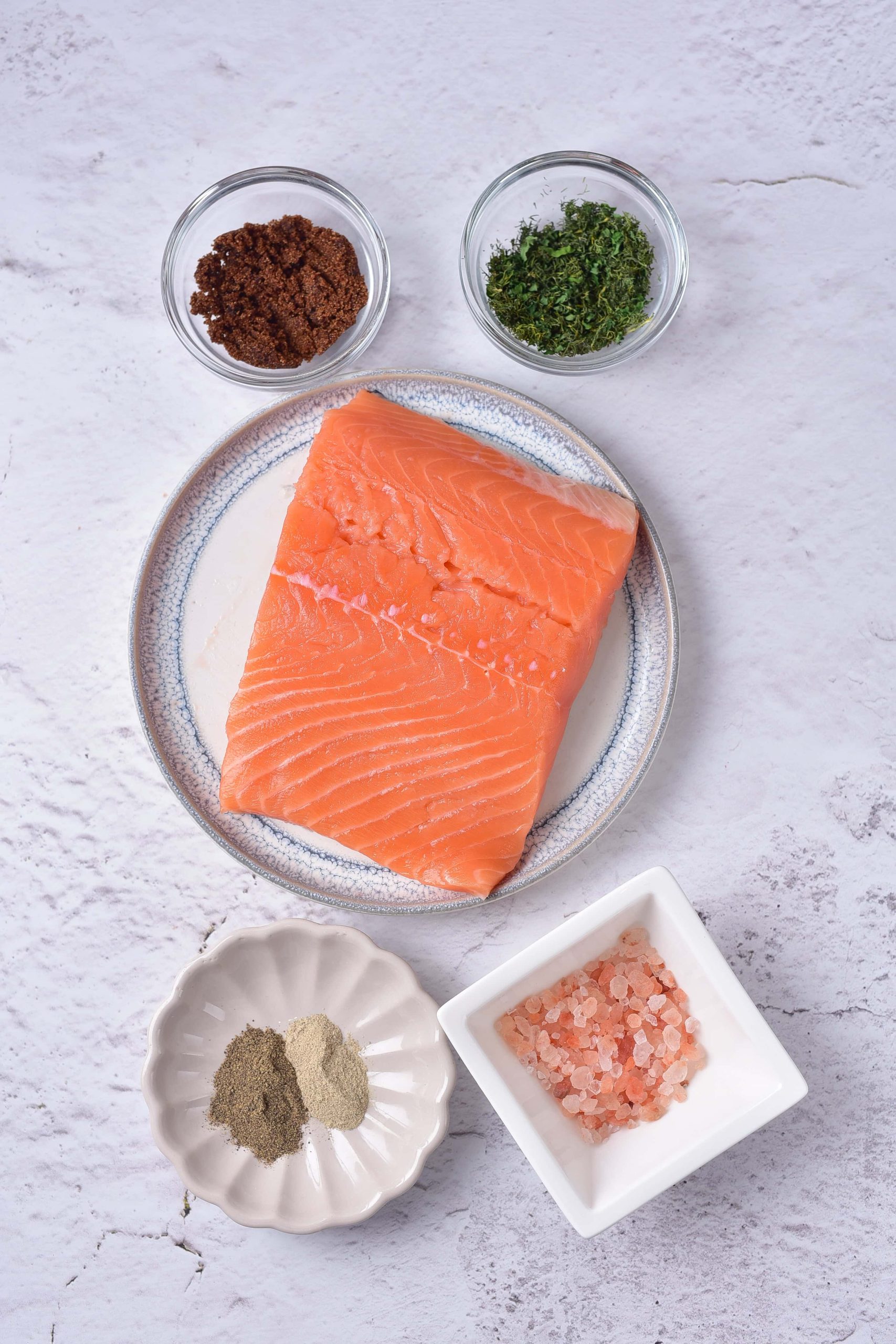 A raw salmon fillet rests on a textured light surface, ready for transformation into gravlax salmon. It’s surrounded by bowls of ground spices, dill, chopped herbs, and pink salt. The setup suggests an easy recipe for culinary adventure.