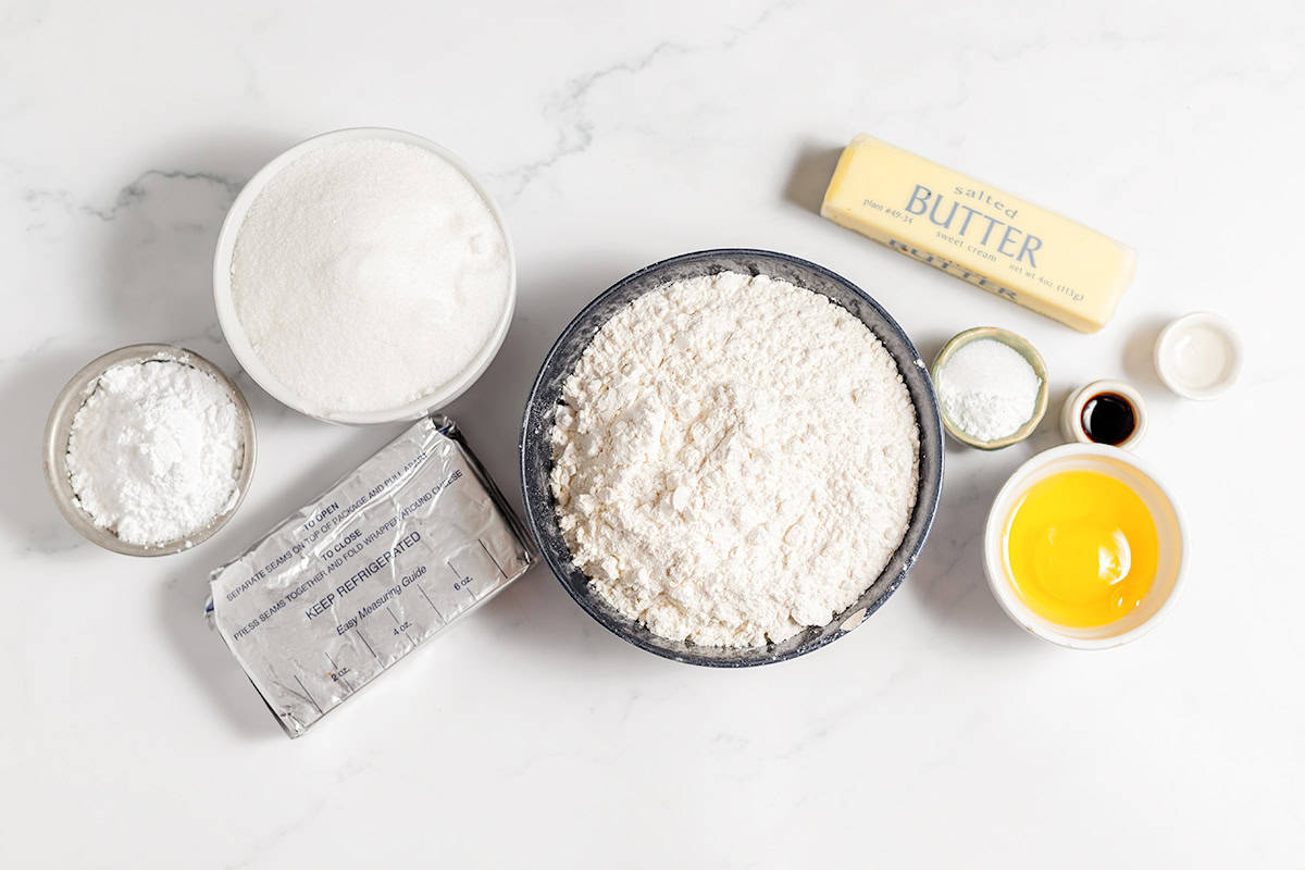 Baking ingredients like flour, sugar, butter, eggs, vanilla extract, baking powder, and salt are arranged on a white surface—perfect for whipping up a batch of gooey butter cookies.