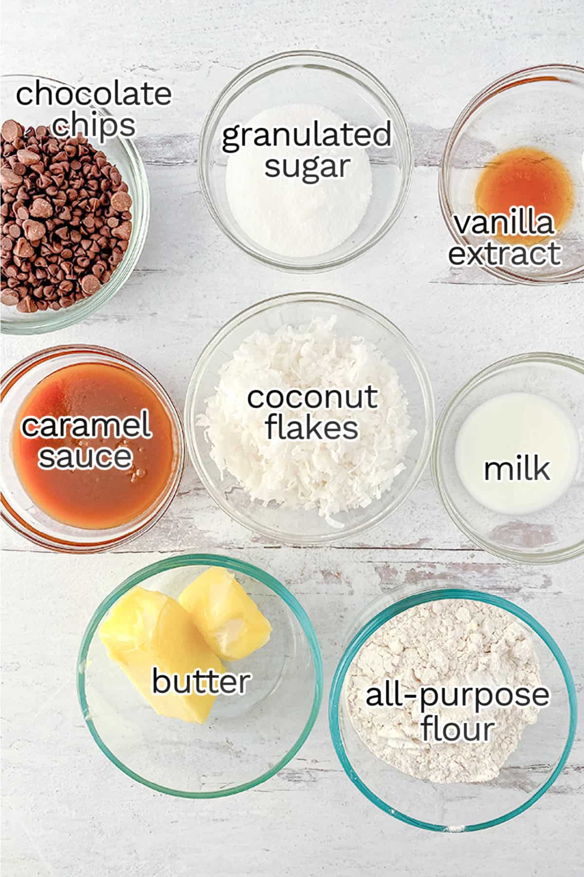 Ingredients for homemade Samoas are displayed in small bowls: chocolate chips, granulated sugar, vanilla extract, caramel sauce, coconut flakes, milk, butter, and all-purpose flour on a white surface.