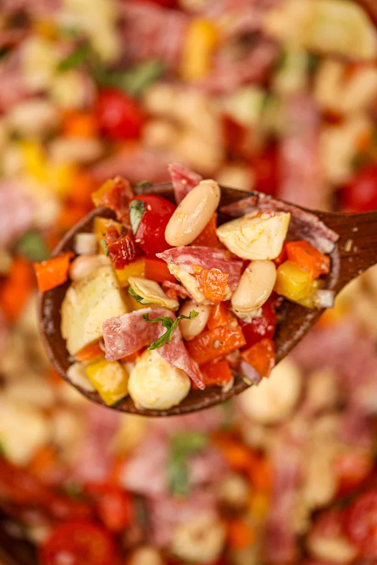 A wooden spoon holding up a small portion of white bean salad with salami and sun-dried tomatoes.