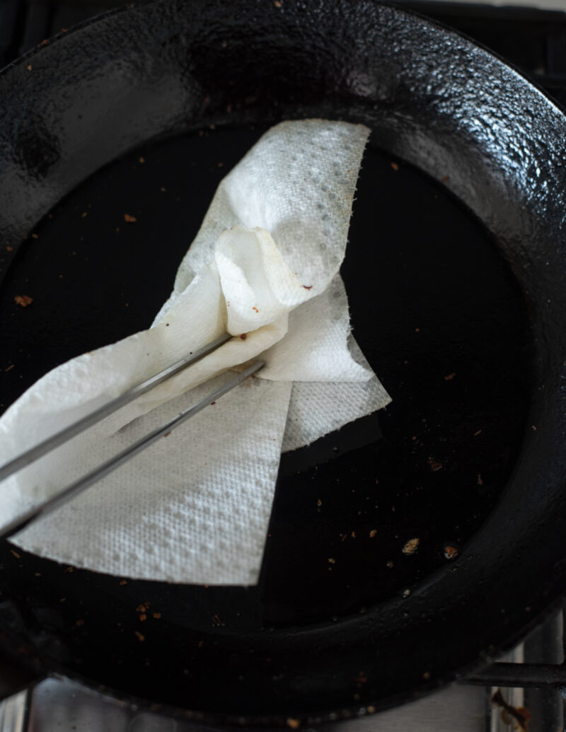 Kitchen tongs holding paper towel to wipe excess oil from skillet