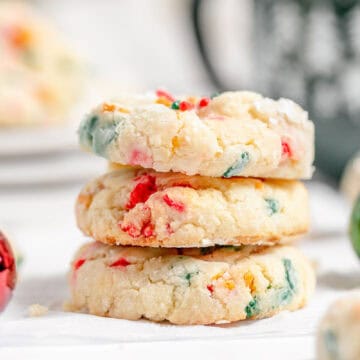 Three colorful gooey butter sprinkle cookies stacked on a white surface, with a blurred mug and additional cookies in the background.
