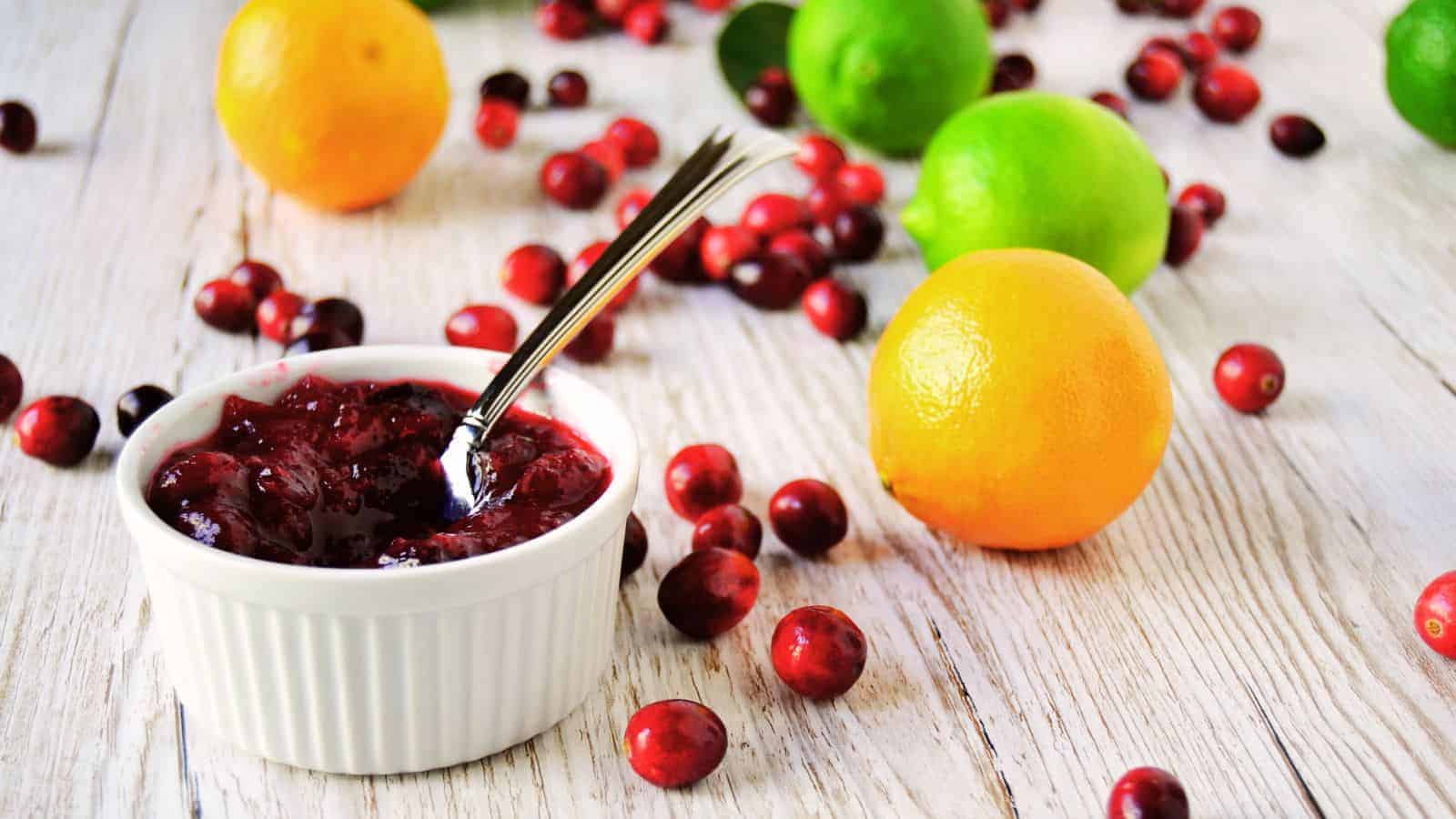 A white ramekin filled with red cranberry sauce sits on a white wooden surface, reminiscent of an Auto Draft. A silver spoon is inside the ramekin, while whole cranberries, green limes, and orange tangerines are scattered around.