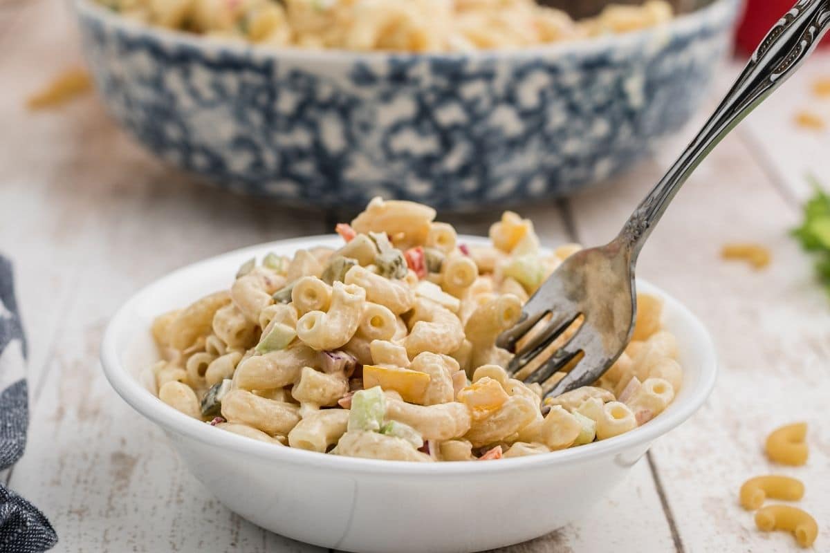 A bowl of creamy macaroni salad with diced vegetables, served with a fork in a white bowl on a light wooden surface.