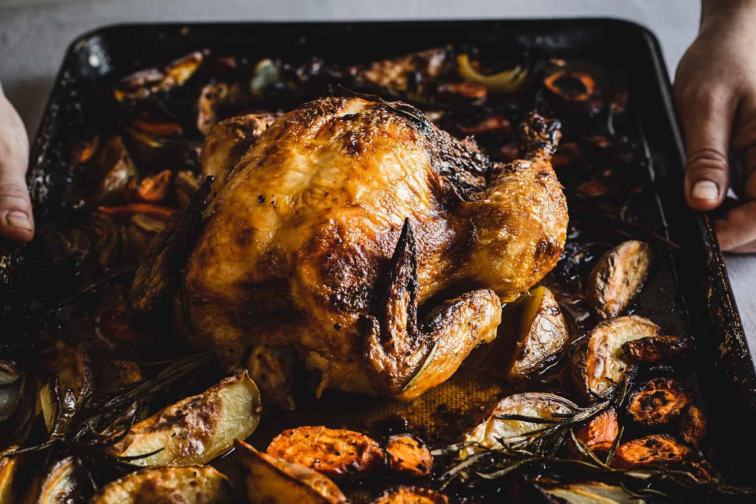 Hands holding paprika chicken and vegetables on a sheet pan.