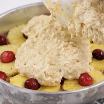 Cake batter being poured on top of pineapples and cherries. 
