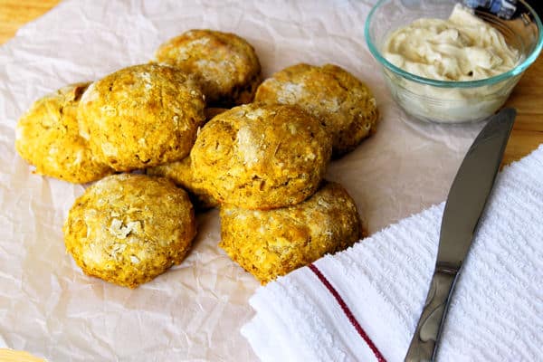 Pumpkin Biscuits stacked on top of each other on a piece of parchment with maple butter, a knife, and a dish towel to the right