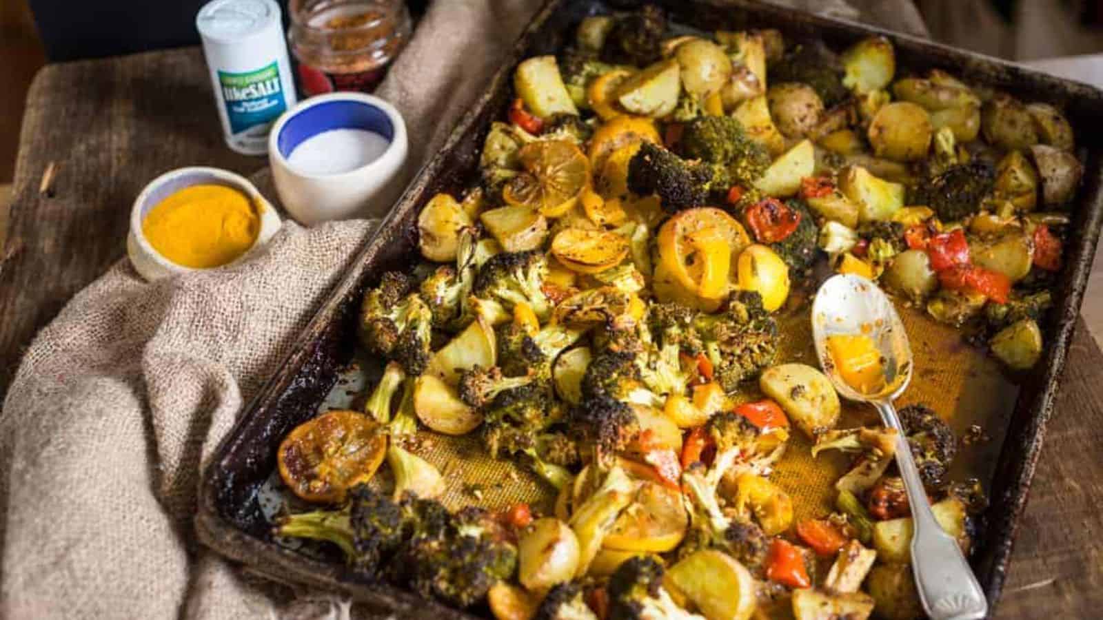 A baking tray with roasted vegetables including broccoli, potatoes, bell peppers, and lemon slices on a wooden table. Nearby, there are containers of spices and a spoon resting on the tray.