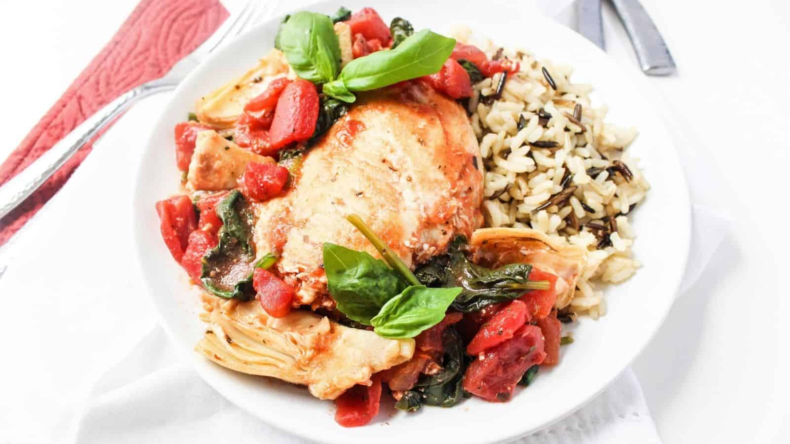 A white plate with chicken, stewed tomatoes, artichokes, sauteed spinach, and wild rice.