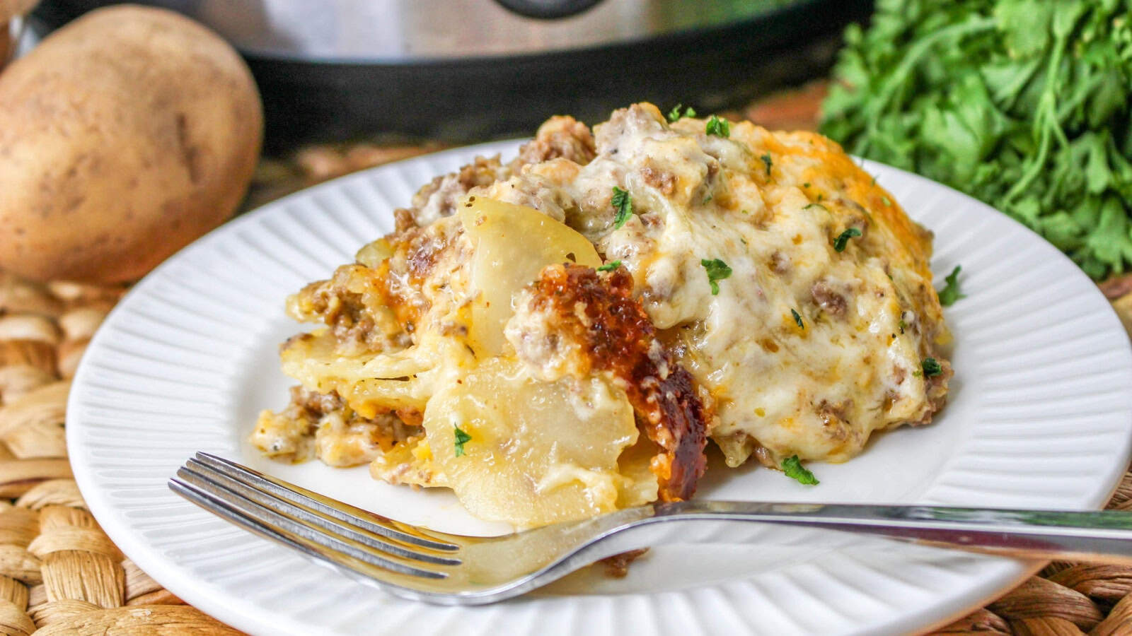 A plate of cheesy beef and potato casserole garnished with herbs, served with a fork. A potato, greenery, and a slow cooker are visible in the background.