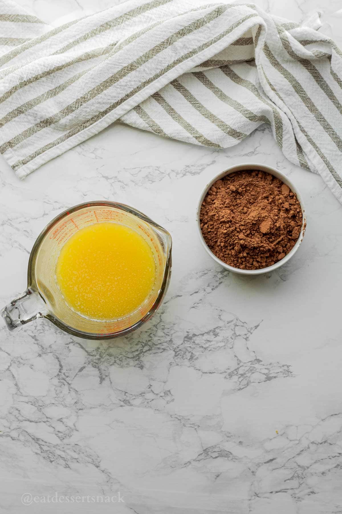 Melted butter and white bowl of cocoa powder on marble countertop.