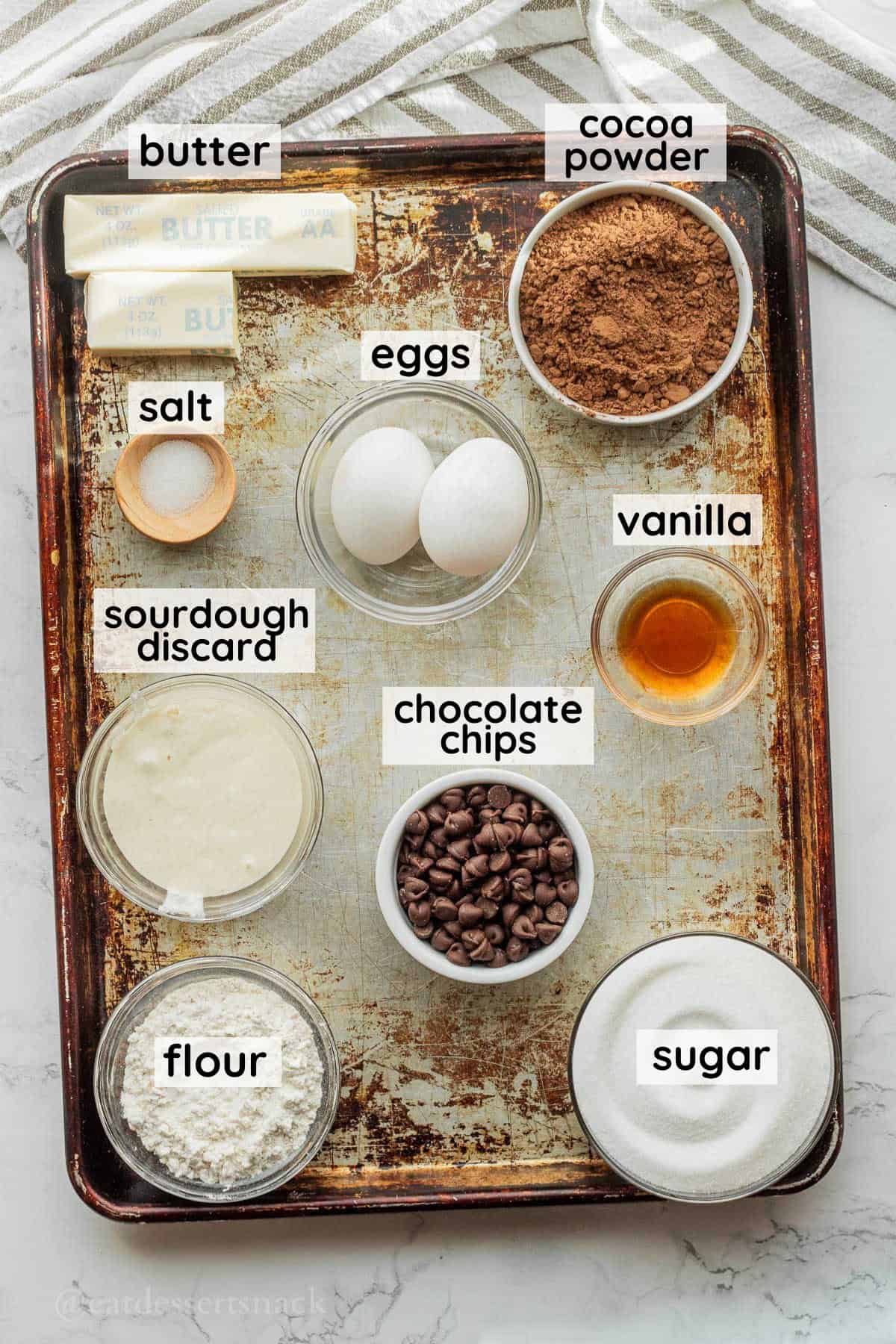 Labeled ingredients for sourdough discard brownies in glass bowls on metal baking sheet.