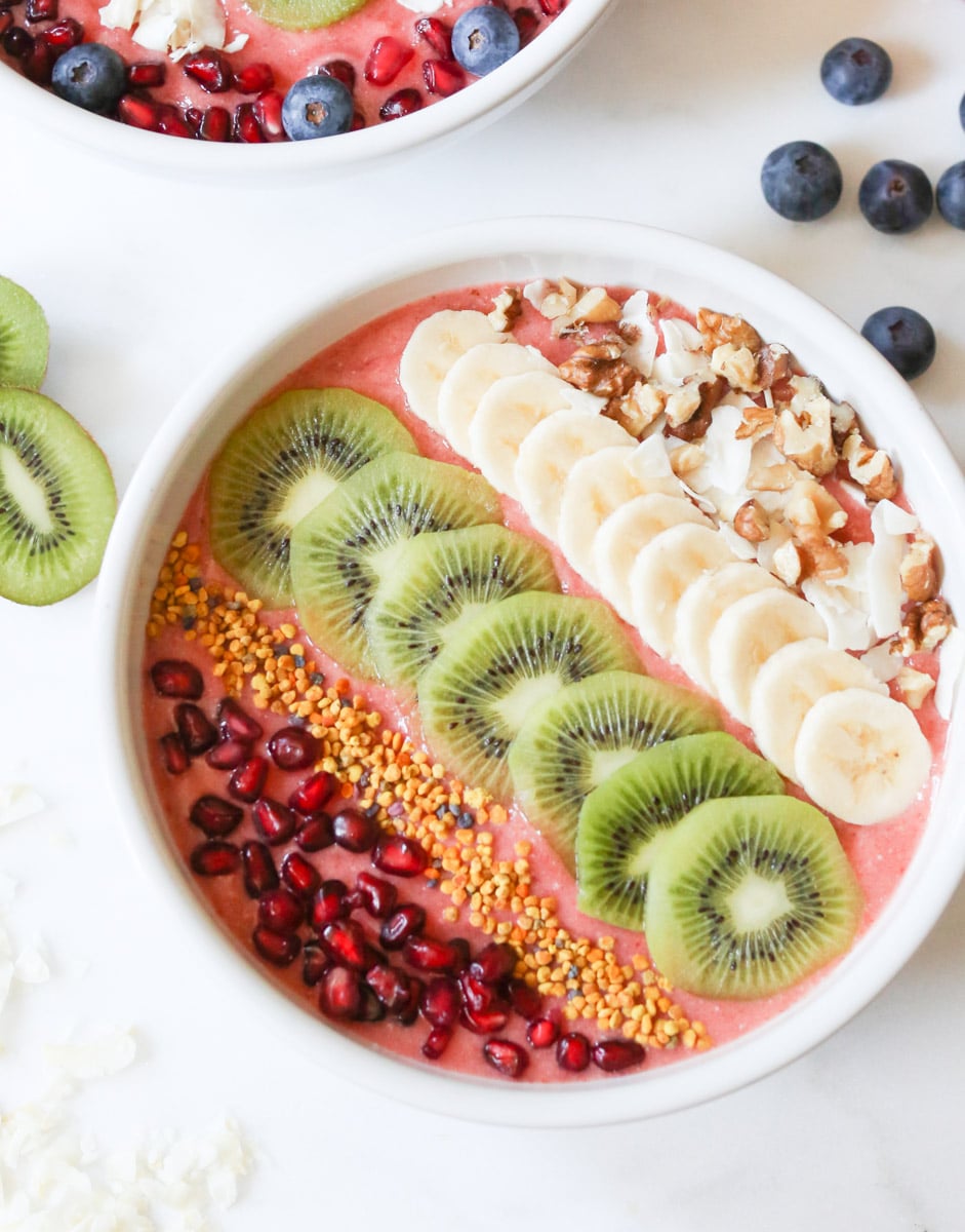 bowl of strawberry kombucha smoothie bowl topped with lines of pomegranate,  bee pollen, kiwi slices, banana slices, and shaved coconut.  