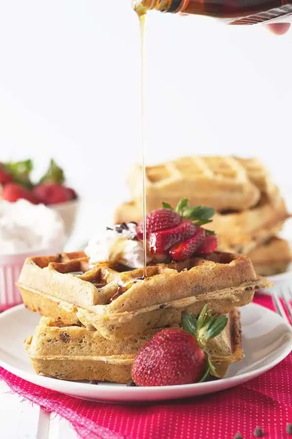 pouring syrup over a stack of 2 strawberry waffles with whipped cream and berries on top