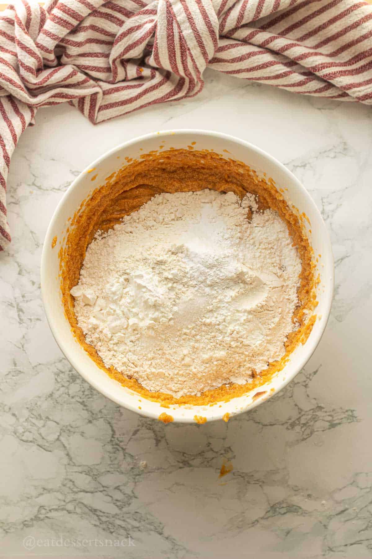 Flour, salt, baking soda, and baking powder on top of pumpkin cookie dough wet ingredients in bowl.