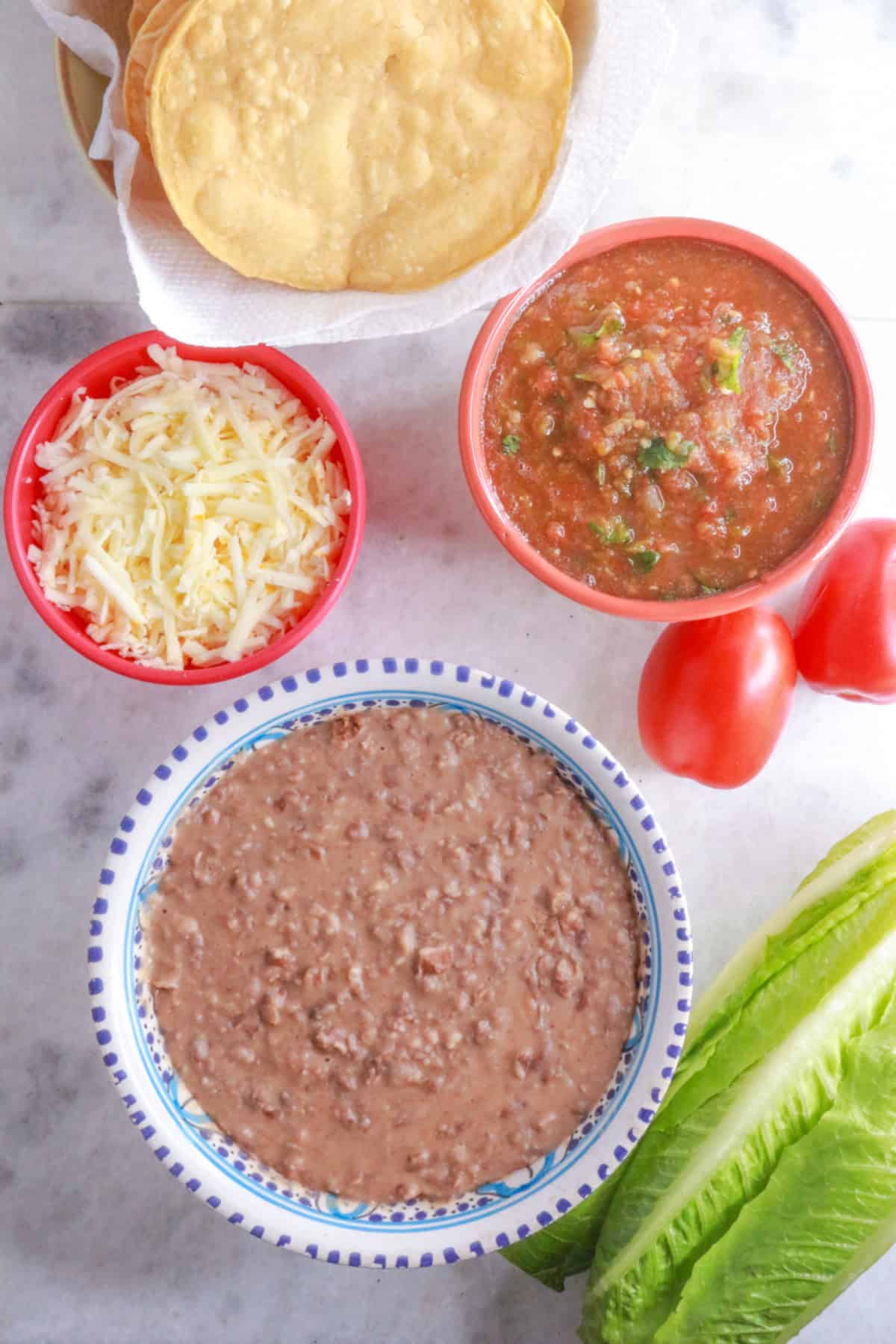 Corn tostadas, a bowl with shredded cheese, a bowl with jalapeño salsa, a bowl with refried beans, 2 roma tomatoes and lettuce